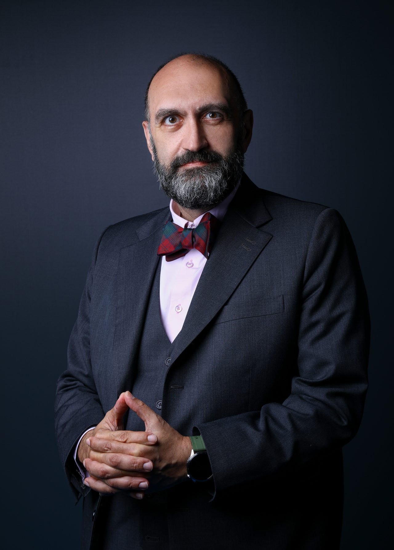 Malek Mroueh in a dark three-piece suit and tartan bow tie, with his hands clasped